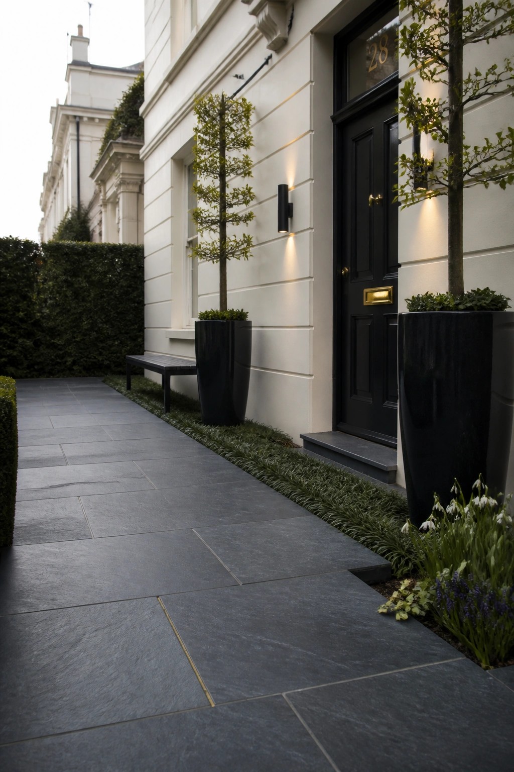 A paved stone walkway leads to a black front door on a white house, with two large black planters holding small trees positioned along the path.