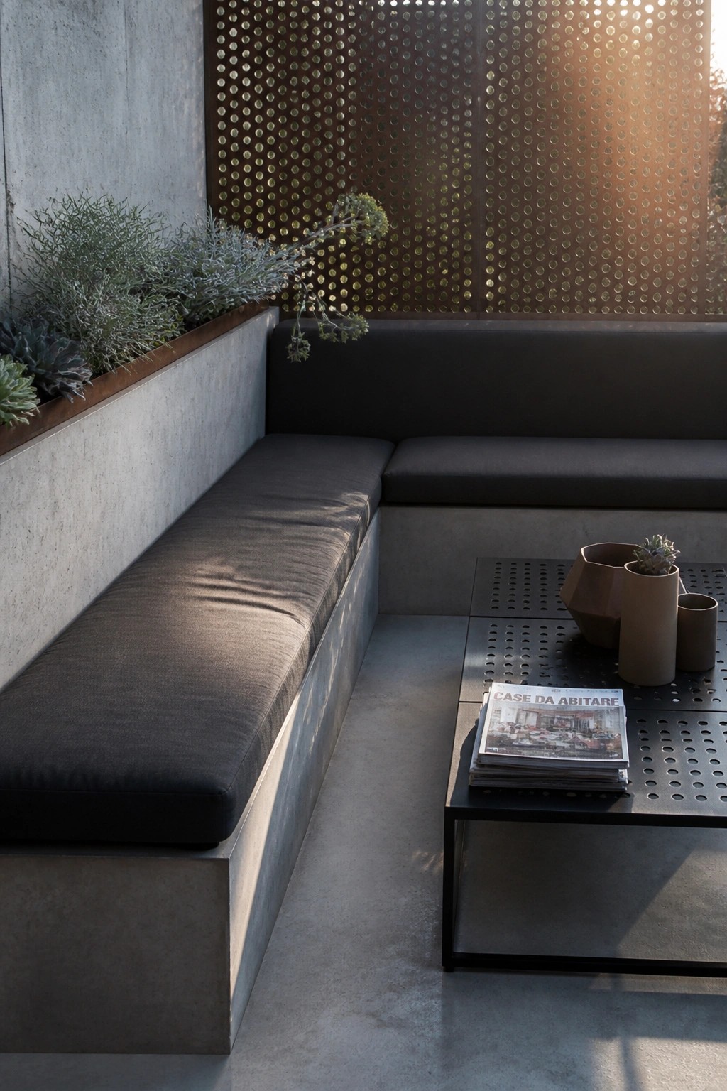 An outdoor corner with a dark cushioned concrete bench, a raised metal-edged planter holding succulents and shrubs, a perforated metal screen behind the seating, and a low black metal table with stacked magazines and ceramic pots.