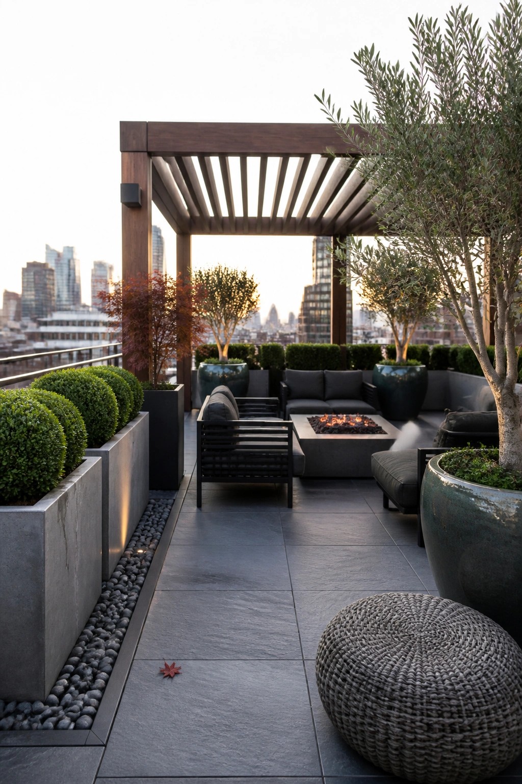 Rooftop terrace with a wooden pergola, gray seating around a fire pit, and large planters holding trees and shrubs overlooking a city skyline.