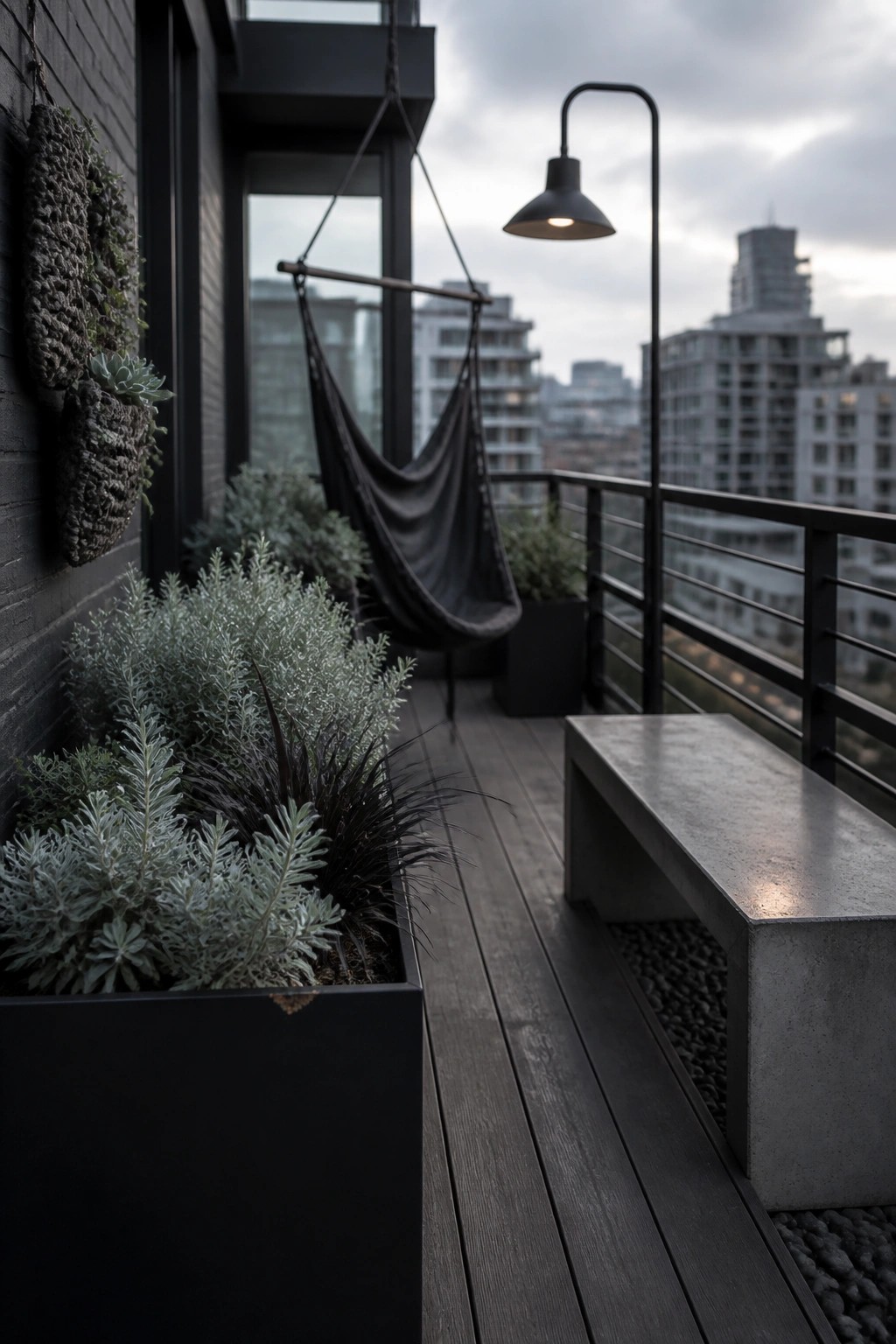 A balcony with dark wood flooring has long black planter boxes along a brick wall holding mixed green and silvery plants, a hanging hammock, and a concrete bench with a city view.