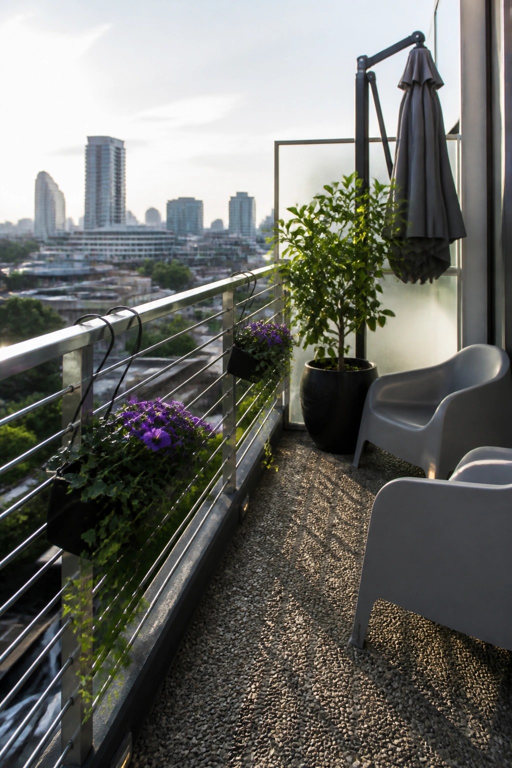 Balcony with metal railing holding two black hanging planters with purple flowers, a large black pot with a tree, and two gray outdoor chairs.