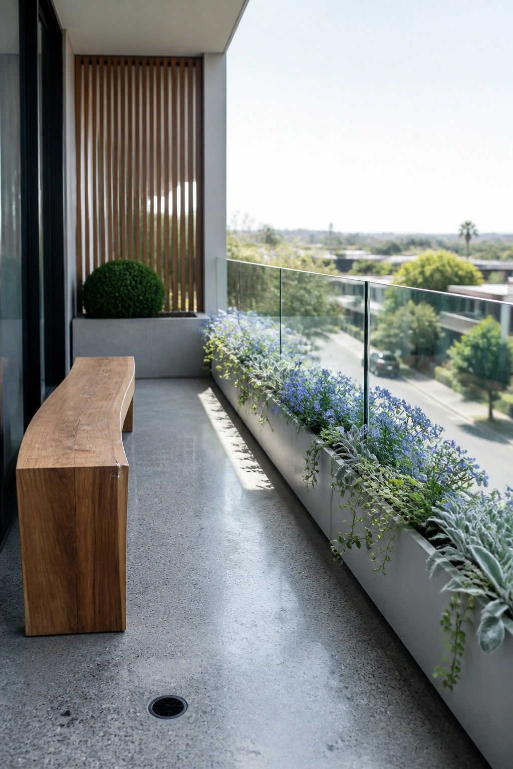 A balcony with a long concrete planter box full of blue flowers and trailing greenery along a glass railing, next to a wooden bench.