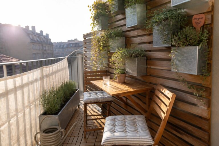 23 Brilliant Small Space Garden Ideas Built Around Vertical Planting and Flexible Containers A small balcony with a wooden slatted wall holding several mounted metal planters, a wooden table with two chairs, striped cushions, and a wheeled metal planter on the floor.