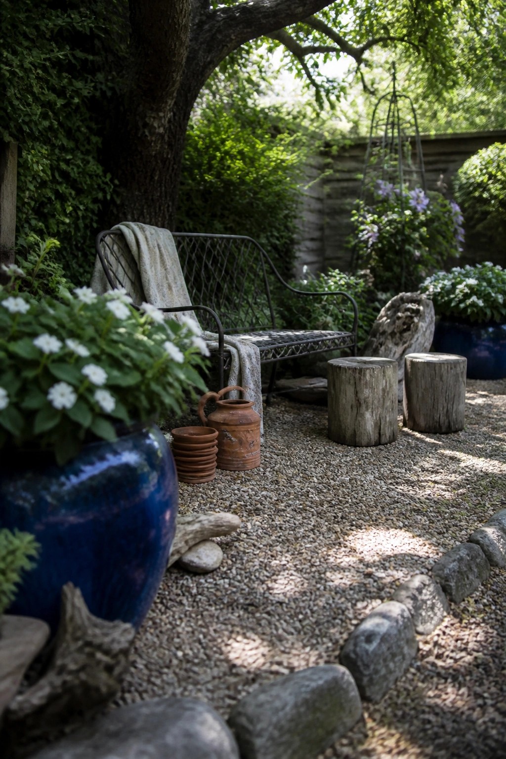 A gravel garden path bordered by large stones includes a metal bench with a throw, a large blue ceramic pot with white flowers, terracotta pots, and two tree-stump stools under leafy trees.