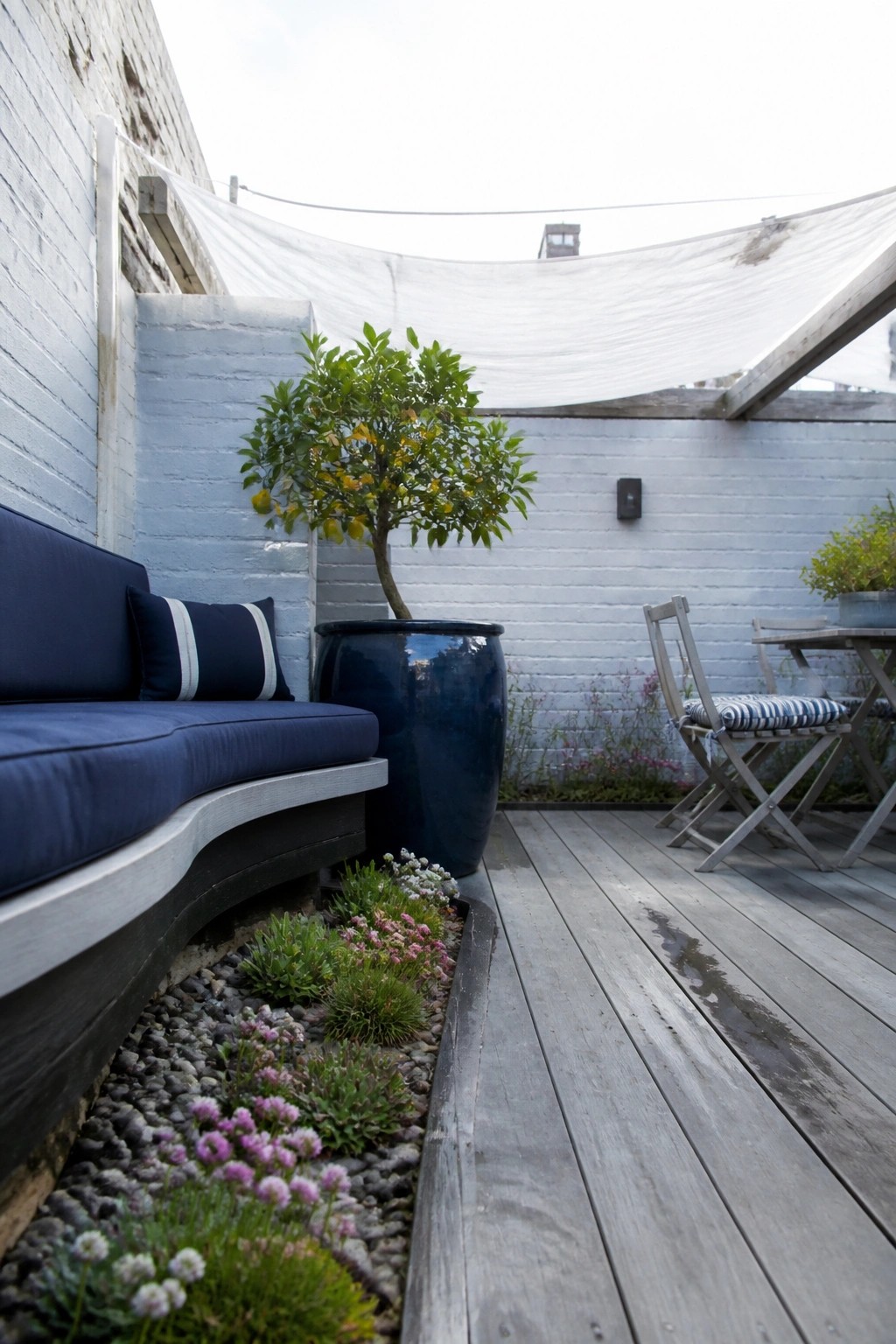 Patio with blue sofa, large potted tree, pebble garden, and wooden chairs.