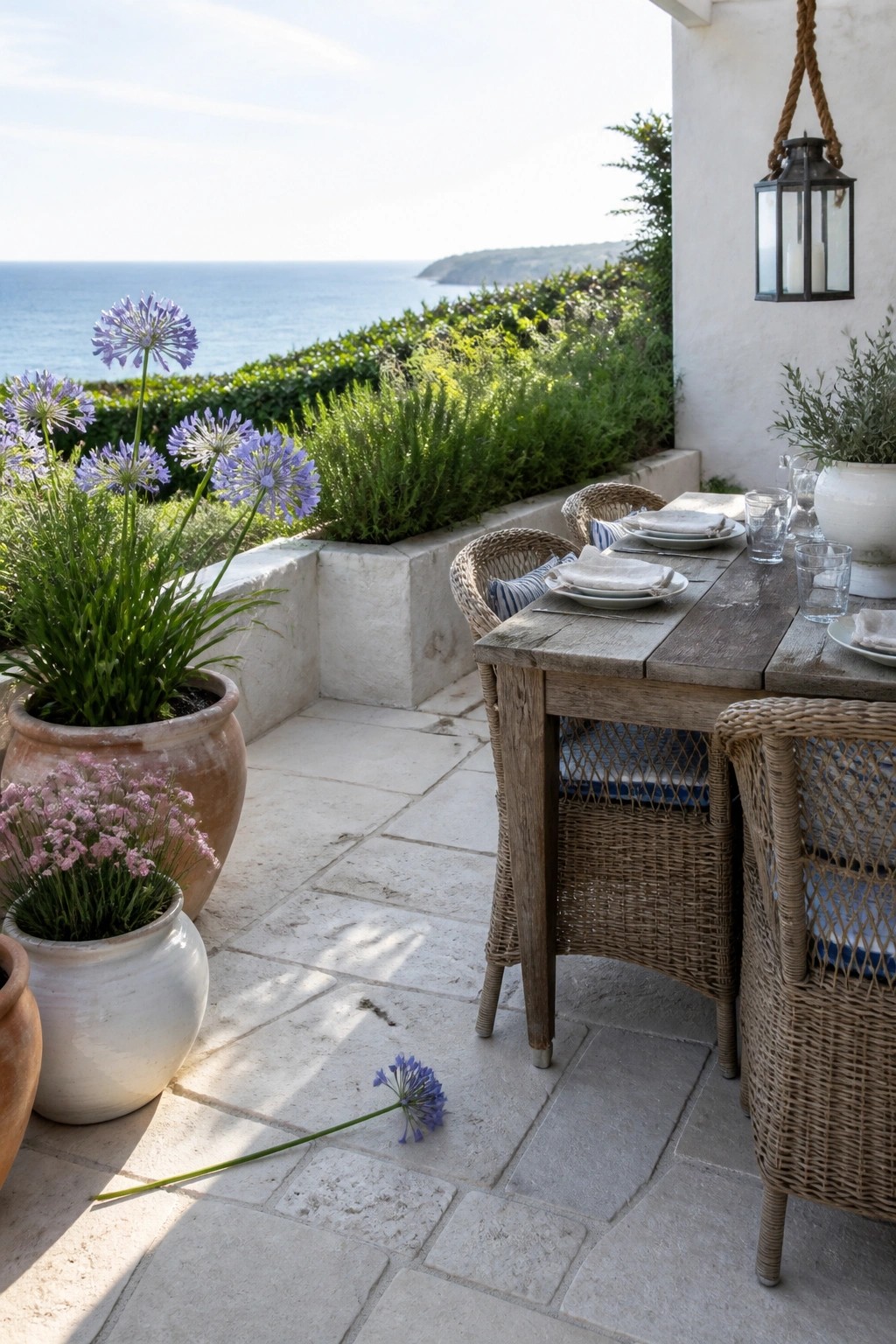A stone paver patio with a wooden dining table, wicker chairs, and several large ceramic pots holding purple and pink flowers, overlooking the ocean.