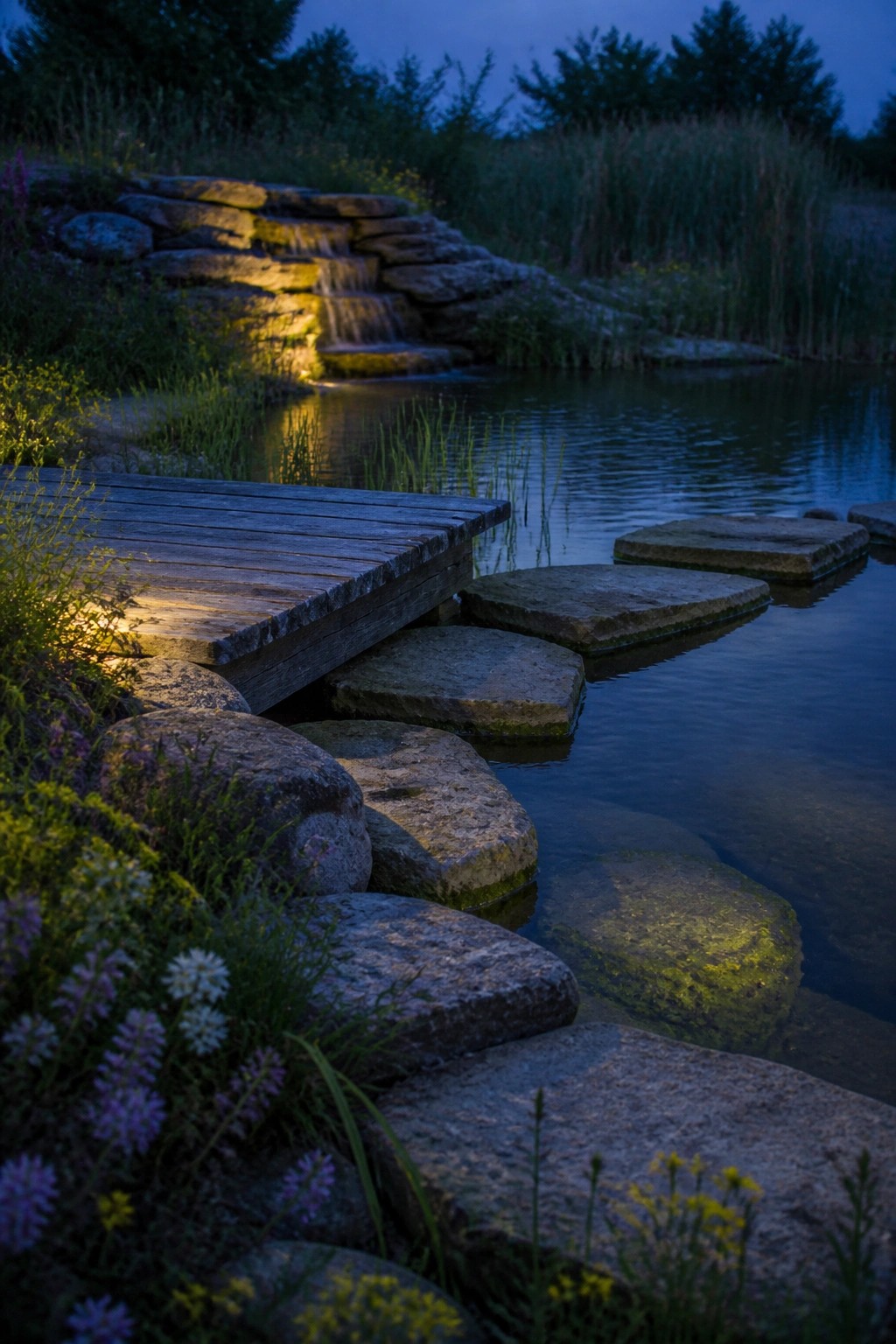 A wooden platform extends beside large flat stepping stones set in a pond, with a stone waterfall and wild plants along the banks at dusk.