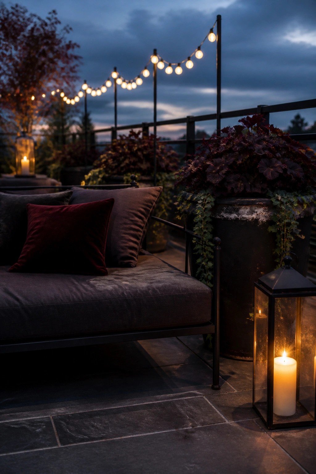 A balcony at dusk with a dark gray daybed, red and gray pillows, large pots of dark foliage, string lights overhead, and glowing lanterns on the stone floor.
