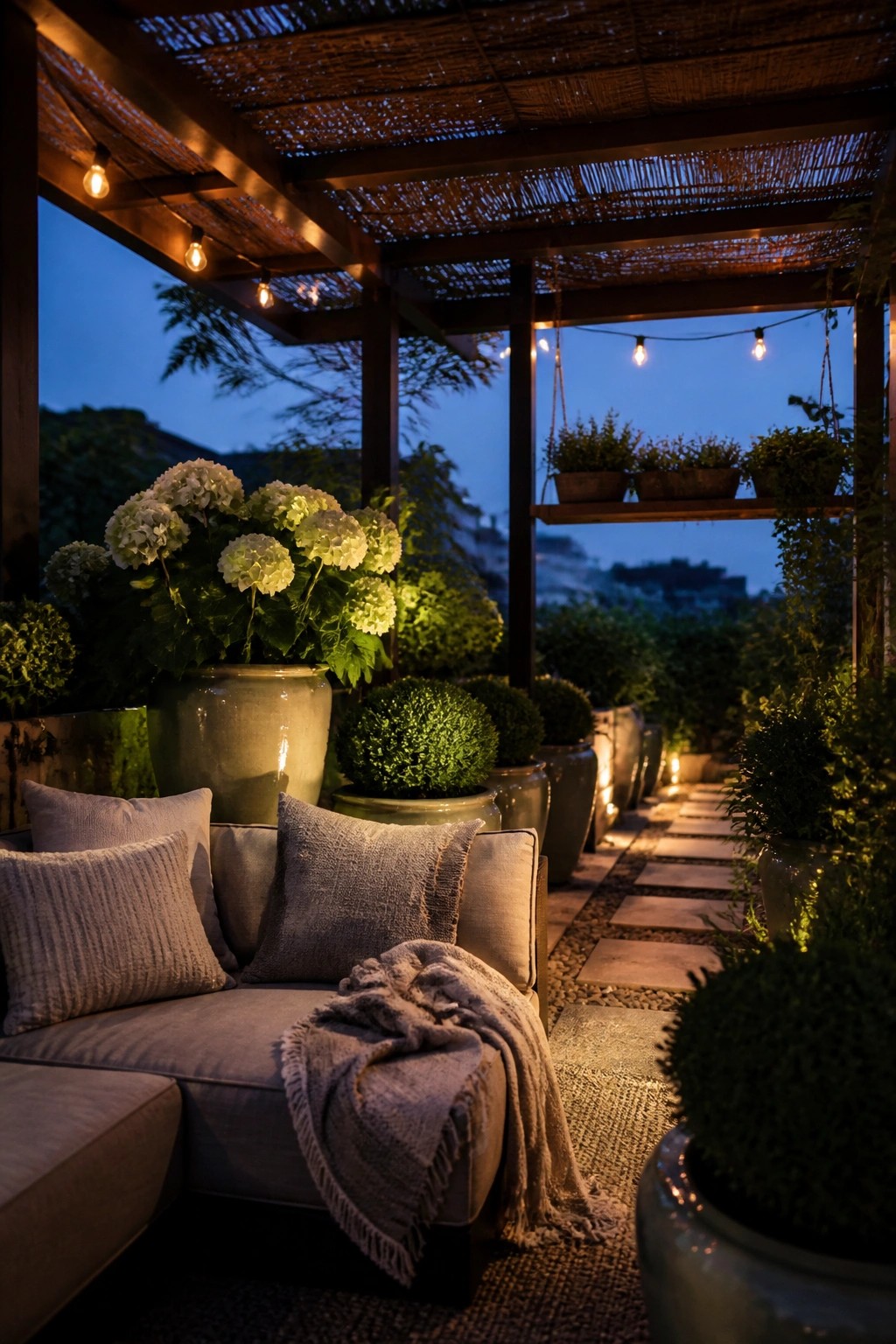 A covered outdoor patio at dusk with string lights under a wooden pergola, a gray sofa with pillows and a throw, and many large potted plants and flowers.
