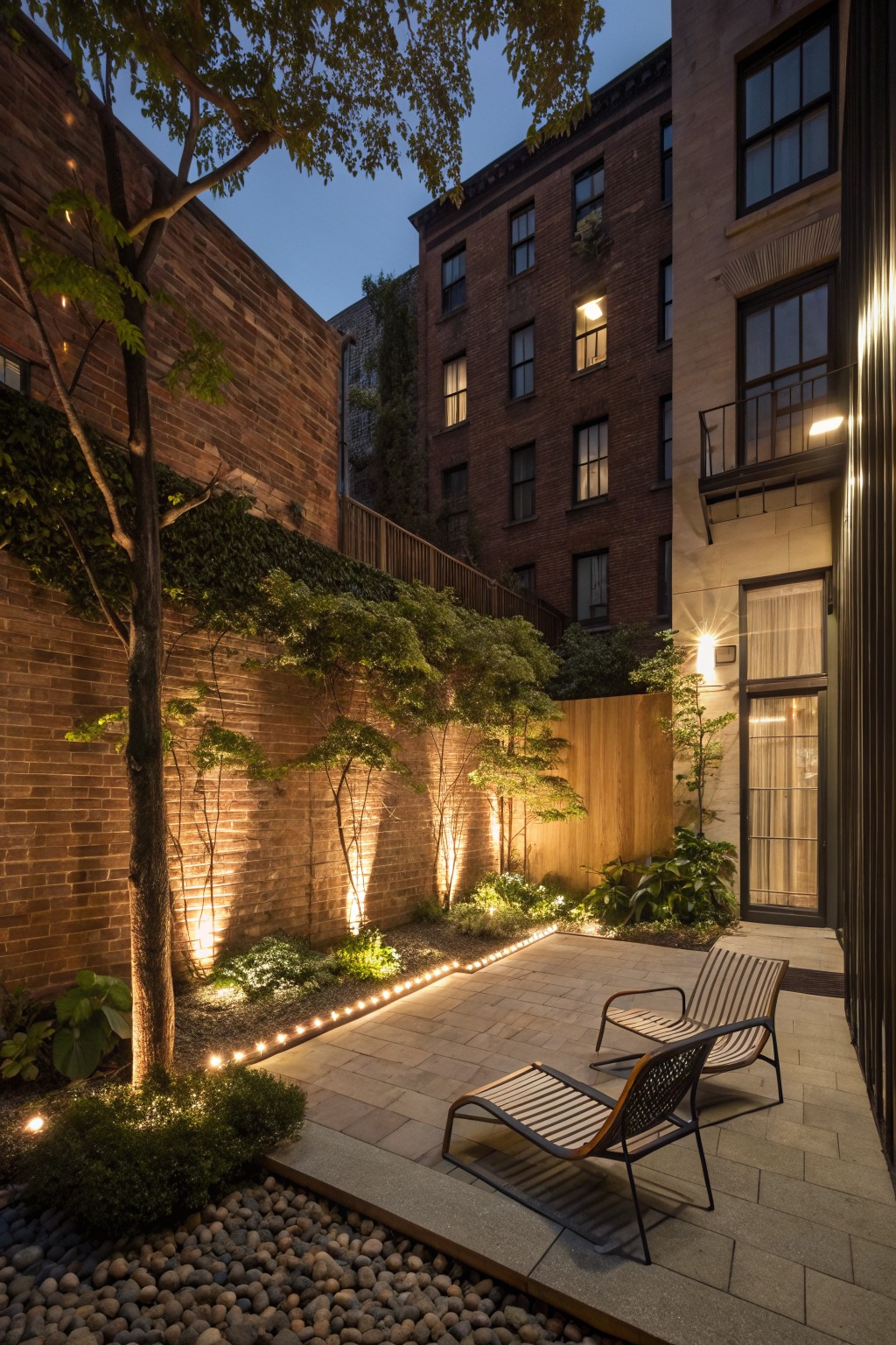 Modern courtyard patio with lounge chairs, lit trees and brick walls at night