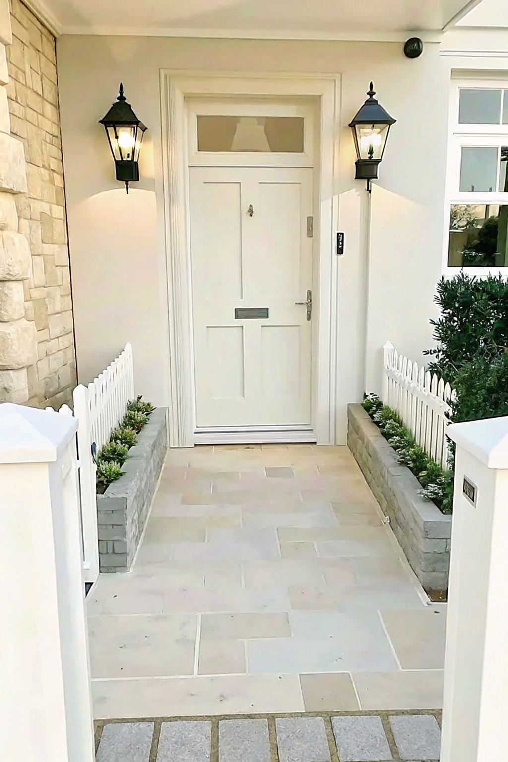 A front entry with a light-colored stone path leading to a white door, flanked by low planted beds edged with white picket fencing and black wall lanterns.