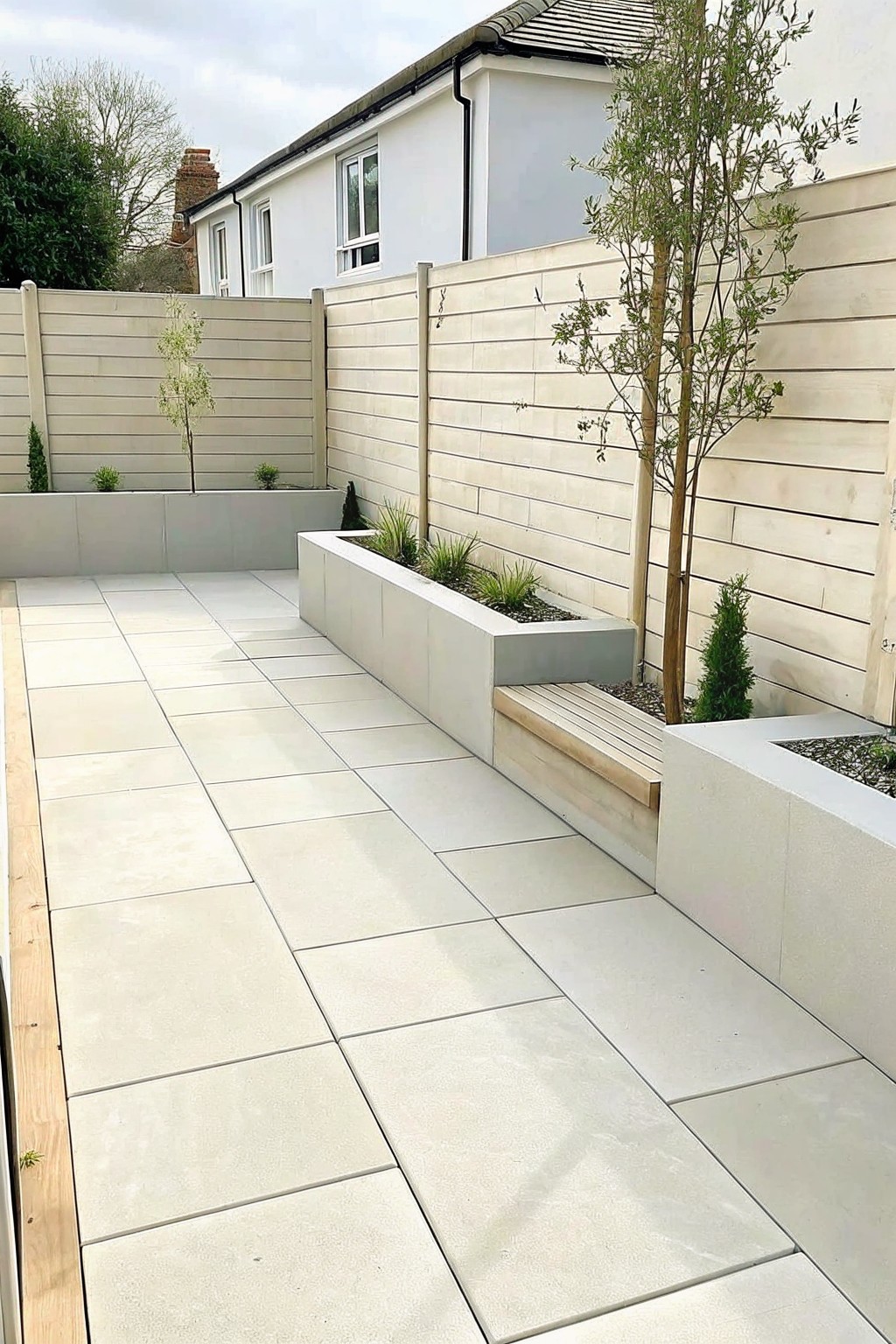 Modern paved courtyard with large light tiles, raised concrete planters holding small trees and shrubs, and a wooden bench built into the side of one planter.