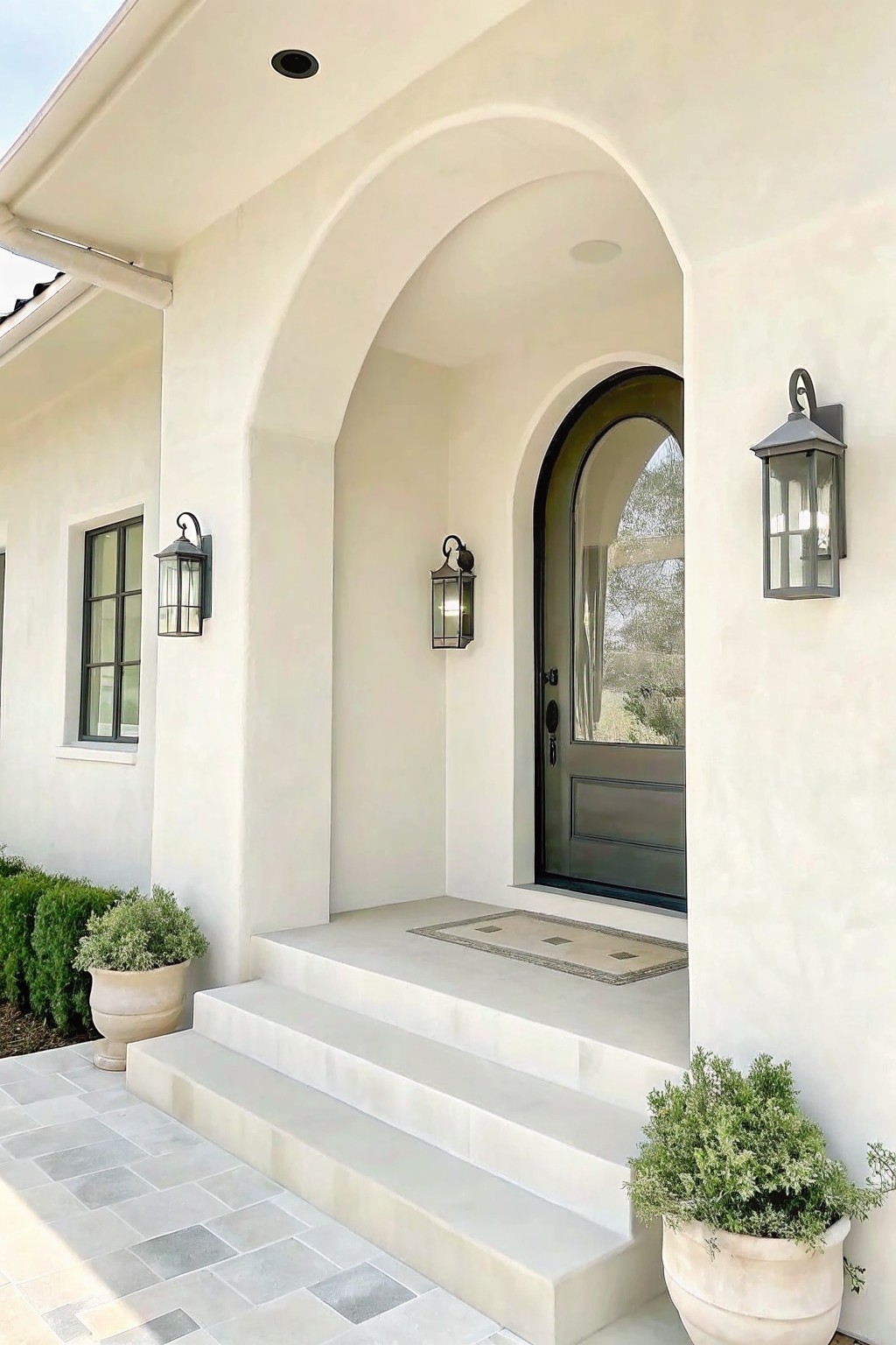 Arched white entryway with dark door, stone steps, wall lanterns, and potted plants