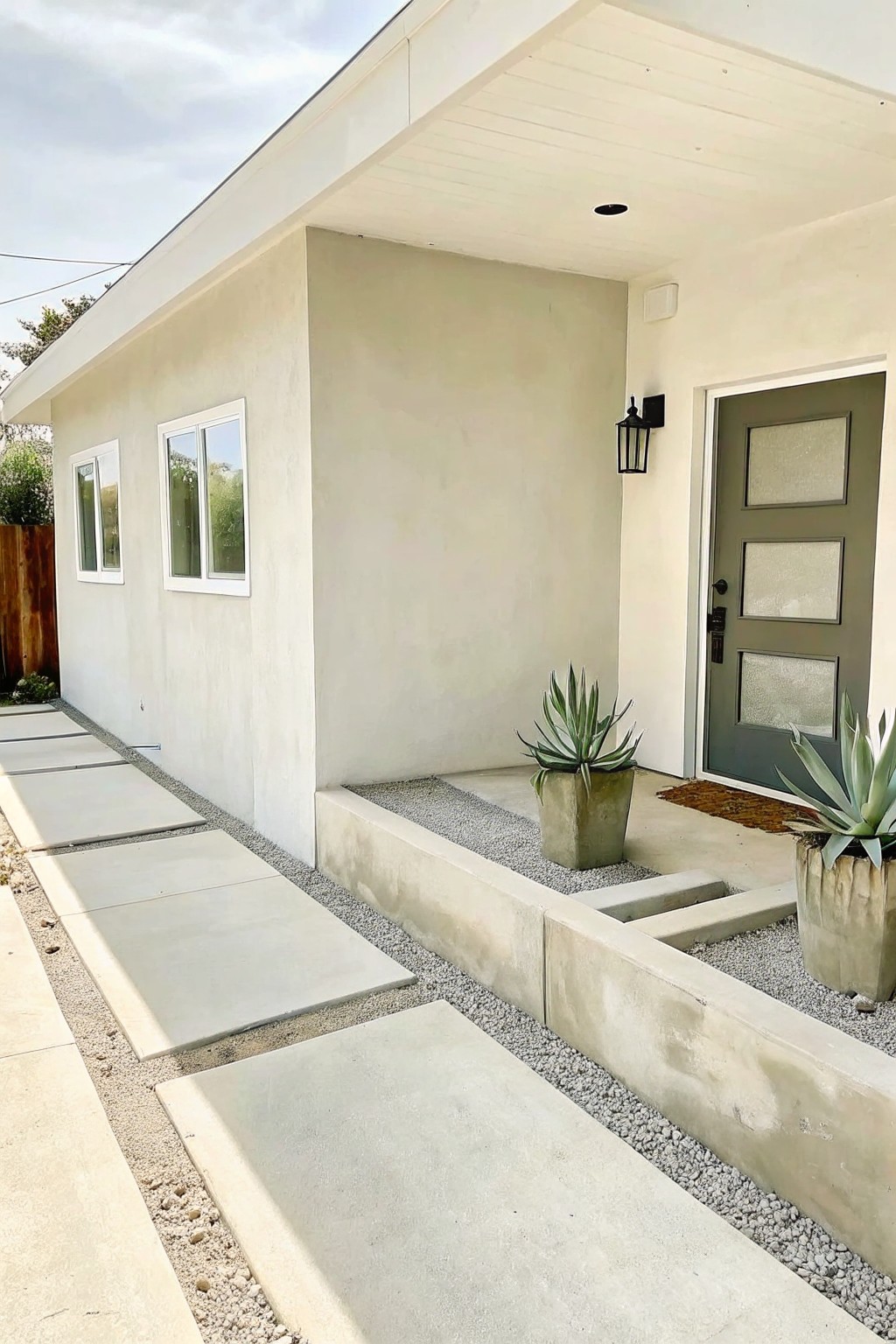 Modern house exterior showing a concrete pathway lined with gravel beds, raised concrete borders, and two large potted agave plants beside a green front door.