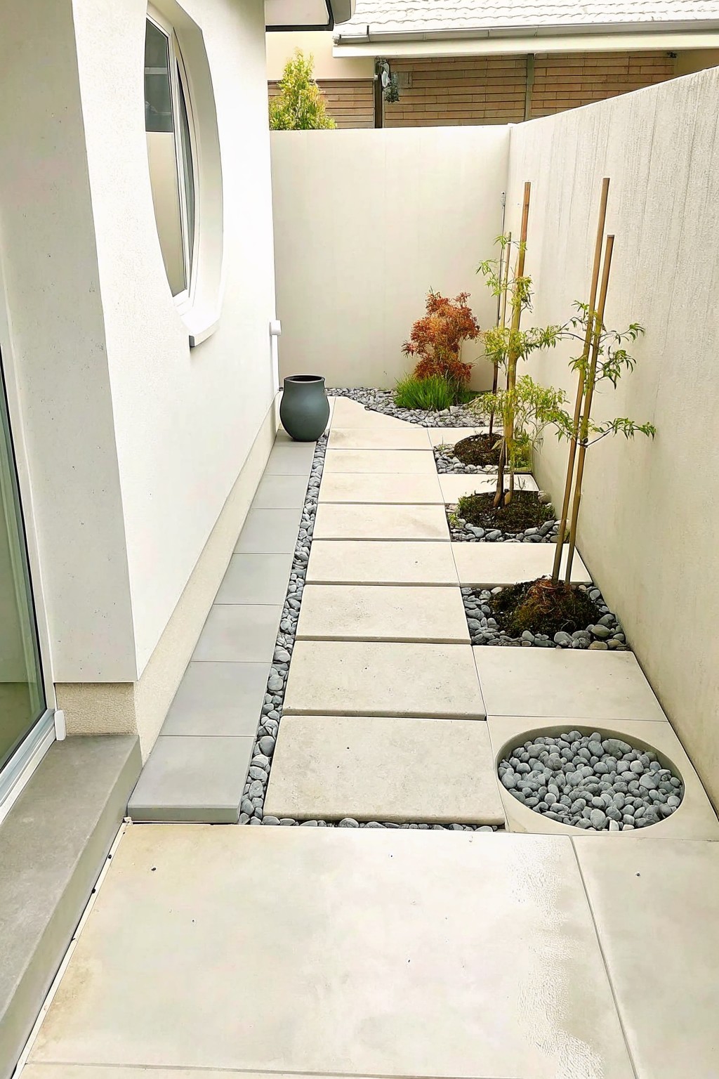 Narrow modern walkway with concrete tiles, pebble borders, staked trees, and white walls.