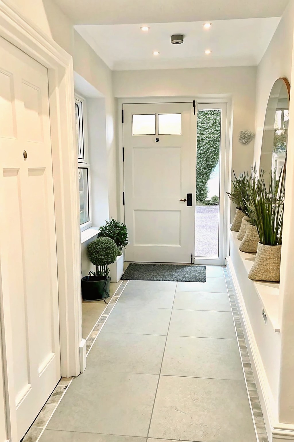 A bright interior entry hallway with light gray floor tiles, white walls and trim, potted plants along both sides, and an open white front door showing a gravel path and greenery outside.