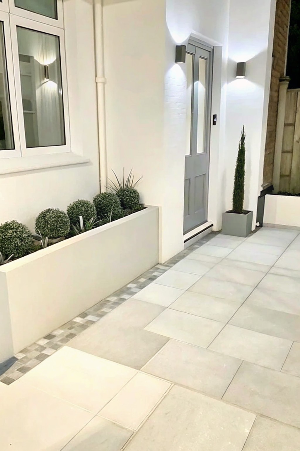 A front walkway with a long white planter box holding rounded green shrubs runs beside light gray tiles leading to a gray door on a white house.