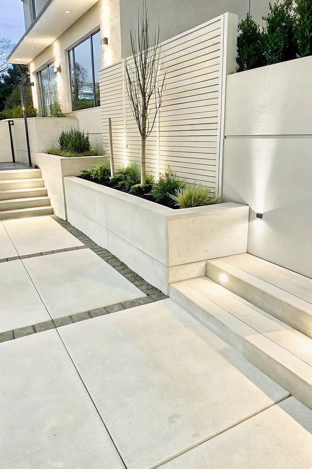 Modern concrete front entry with raised rectangular planters, steps, paving, and mixed shrubs including a small tree.