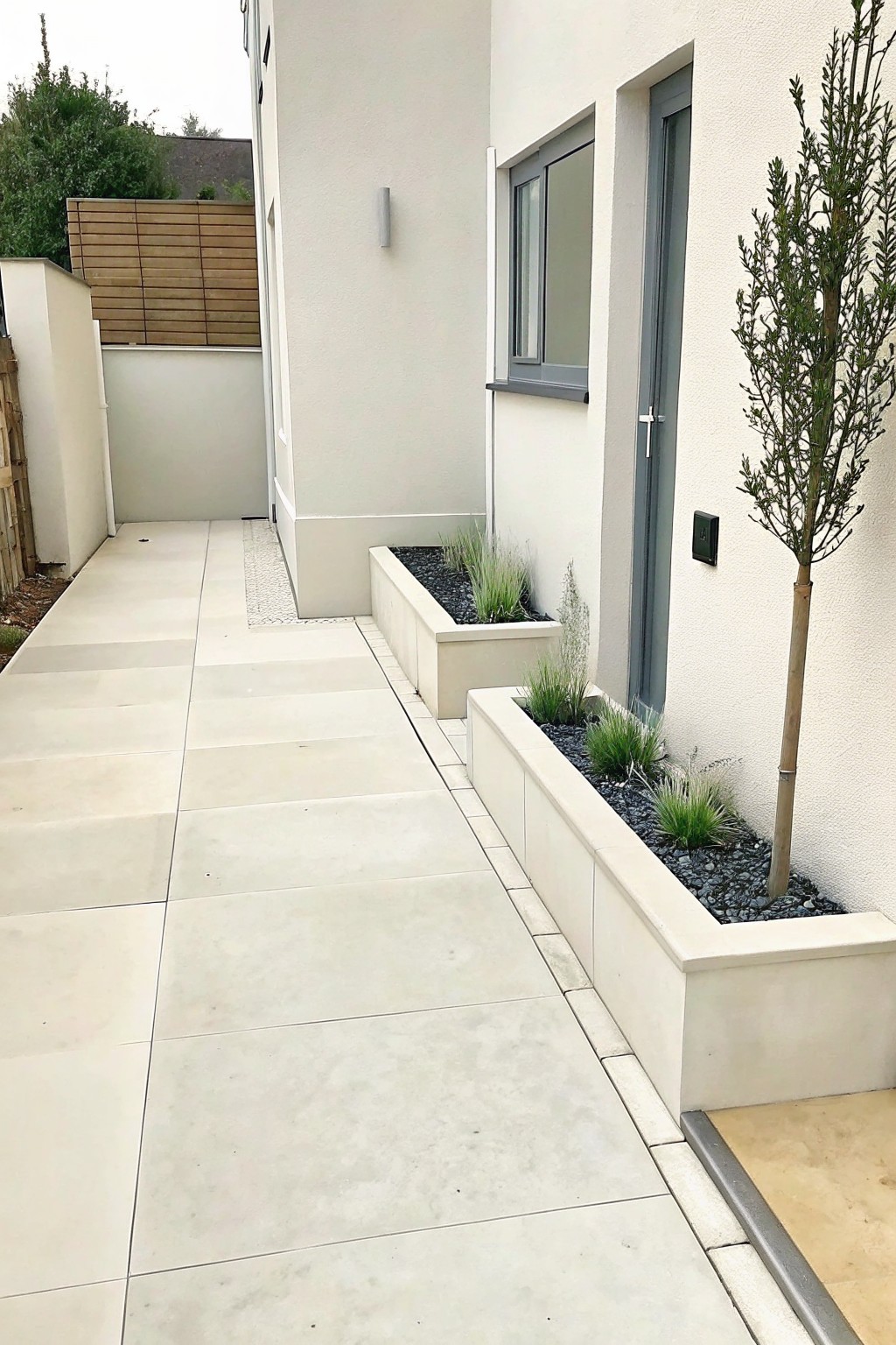 A paved concrete walkway runs beside a modern house, bordered by long raised concrete planters filled with black gravel and ornamental grasses.