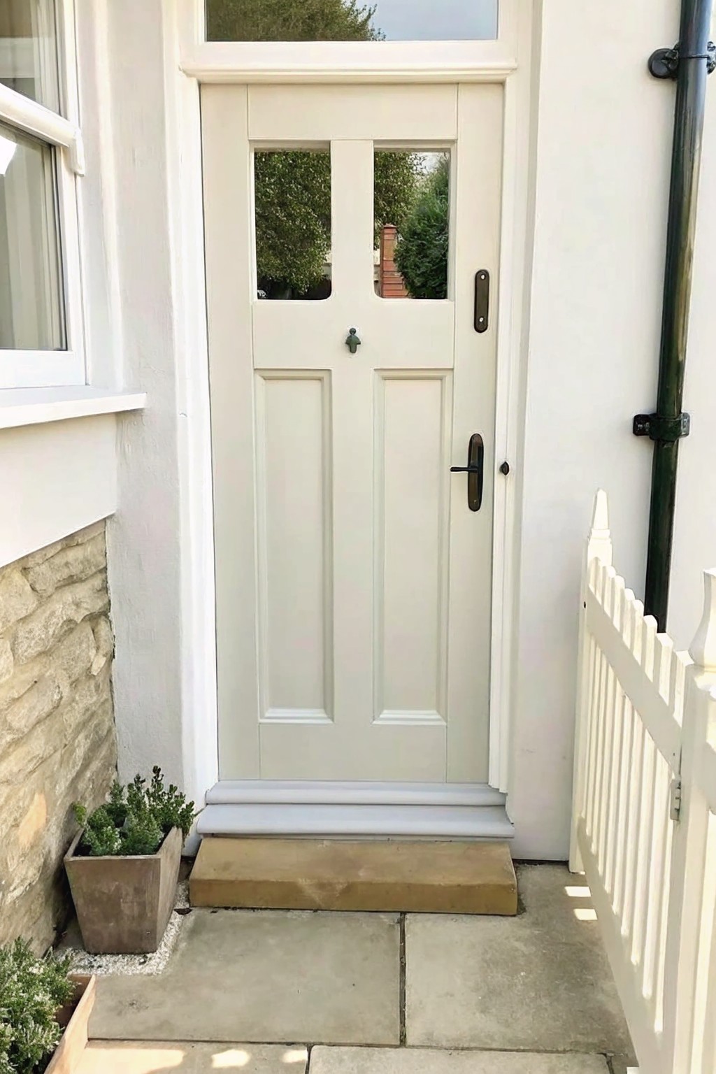 A white paneled front door with gray steps, a wooden planter box holding small green shrubs on the left side, and a white picket fence visible on the right.