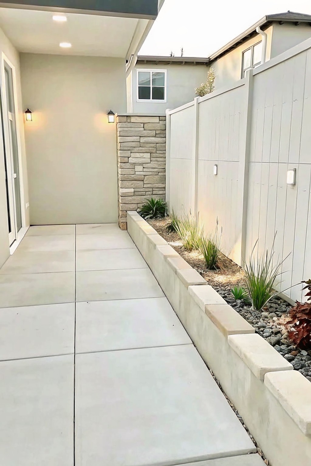 Narrow concrete walkway beside low garden wall, plants, and white fence