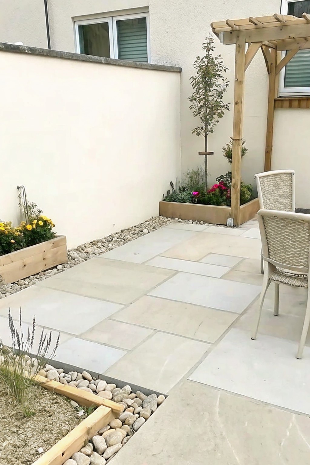 A paved stone patio bordered by gravel strips and wooden raised planters filled with flowers, lavender, and a small tree, with a pergola and chairs visible on the right.