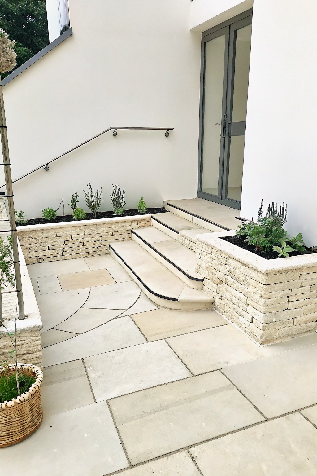 A modern outdoor area with light stone steps, raised stone planters holding green plants, and large beige paving stones on the ground.