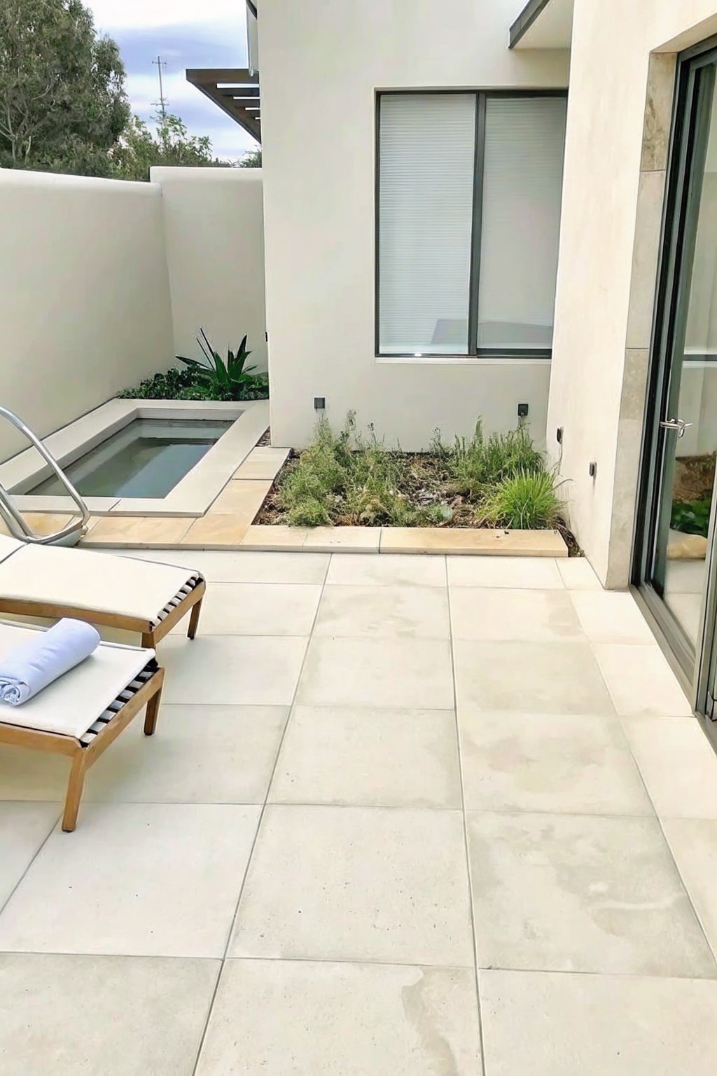 A light-colored tiled patio with a wooden lounge chair, a small rectangular pool, and a narrow rectangular bed of mixed grasses and wildflowers bordering the wall.