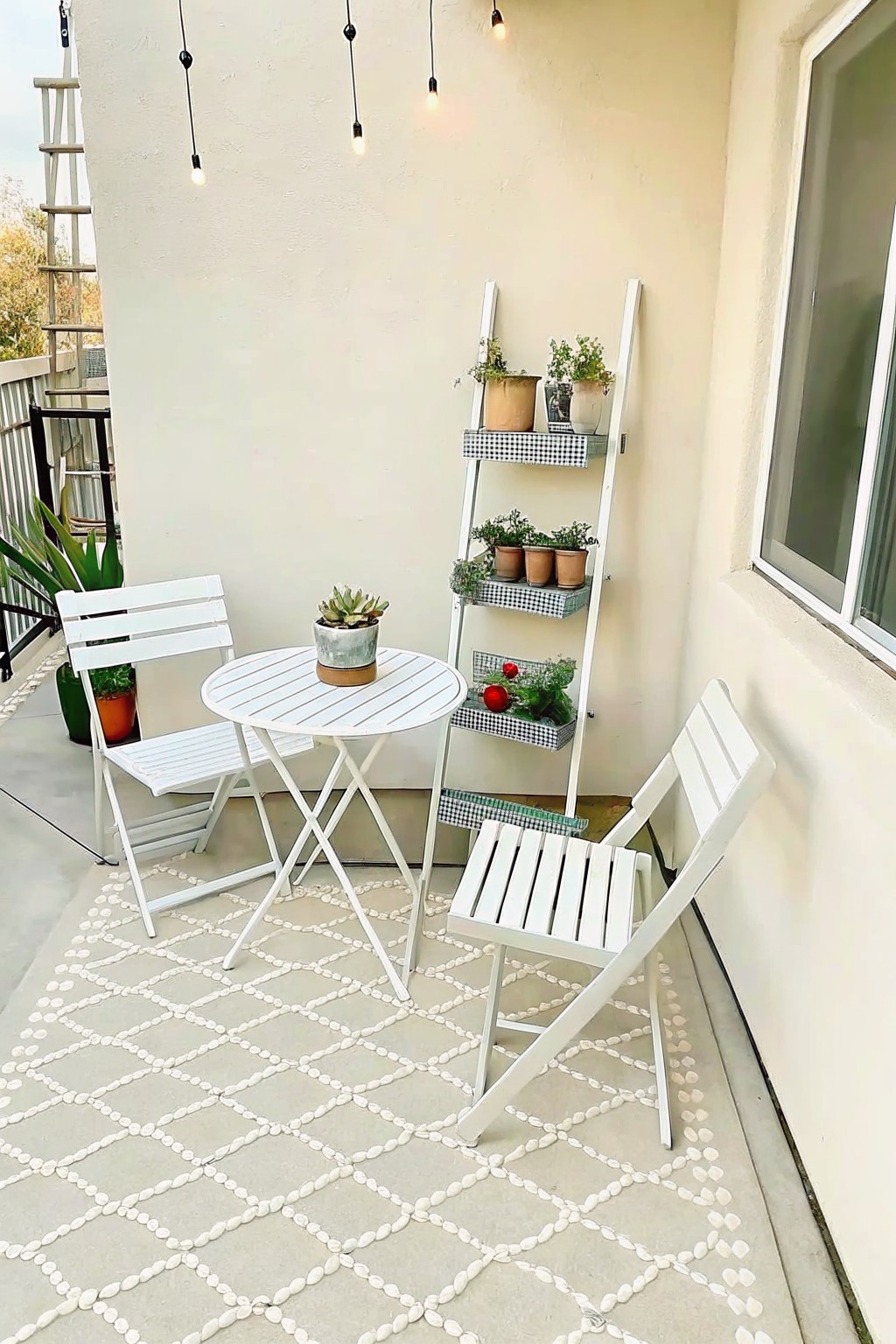 A small balcony with a round white bistro table and two white folding chairs placed on a light geometric outdoor rug, next to a white leaning ladder shelf holding potted plants.