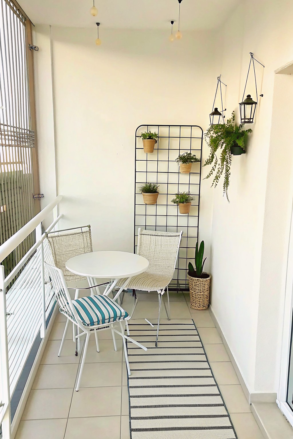 A narrow balcony with a white round table, two woven chairs, a black metal grid holding potted plants, hanging lanterns on the wall, and a striped rug on the tiled floor.