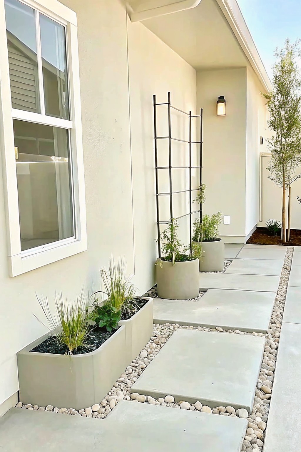 A narrow concrete path beside a house lined with large beige planters, a black metal trellis, and pebble ground cover.