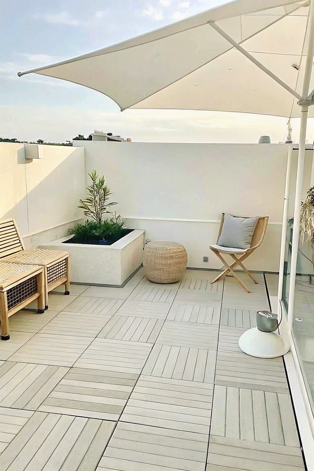 Rooftop terrace covered in light gray modular deck tiles with a large white shade sail, wooden seating, a built-in style planter, and a woven ottoman.