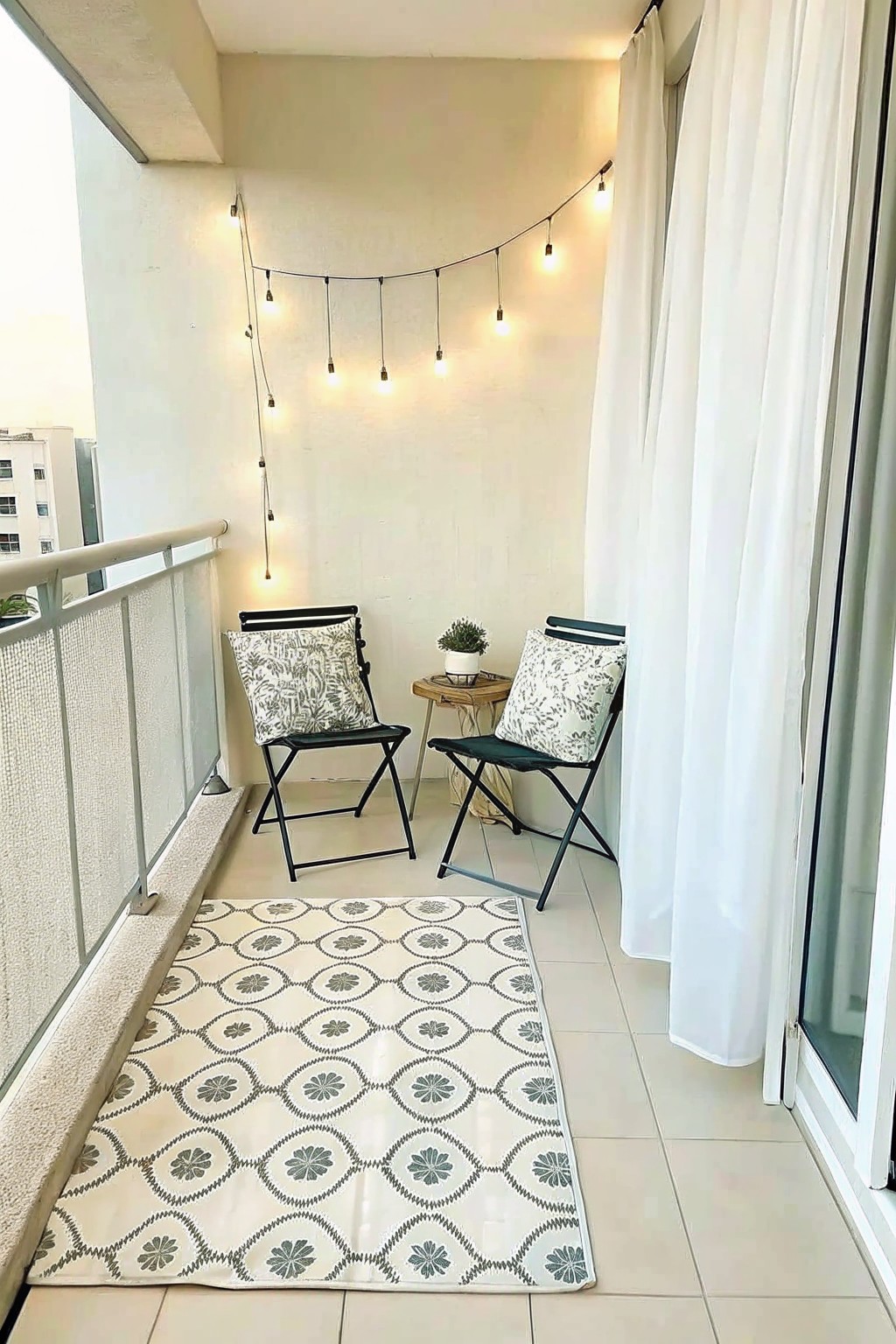 A small balcony with two folding chairs, a patterned rug, a small side table with a plant, and string lights hung along the back wall.