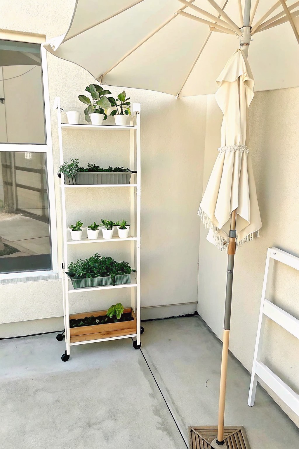 A white rolling shelving unit with multiple levels of potted plants sits on a concrete patio beside a closed beige patio umbrella.