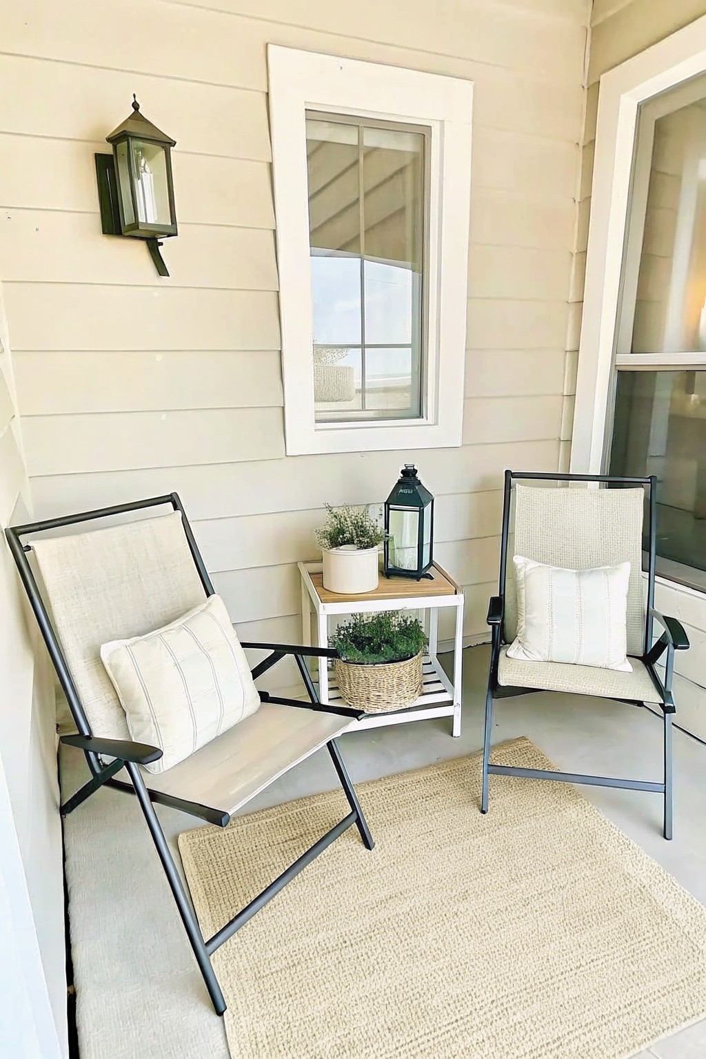 Two cushioned chairs with pillows on a porch rug beside a plant-filled table