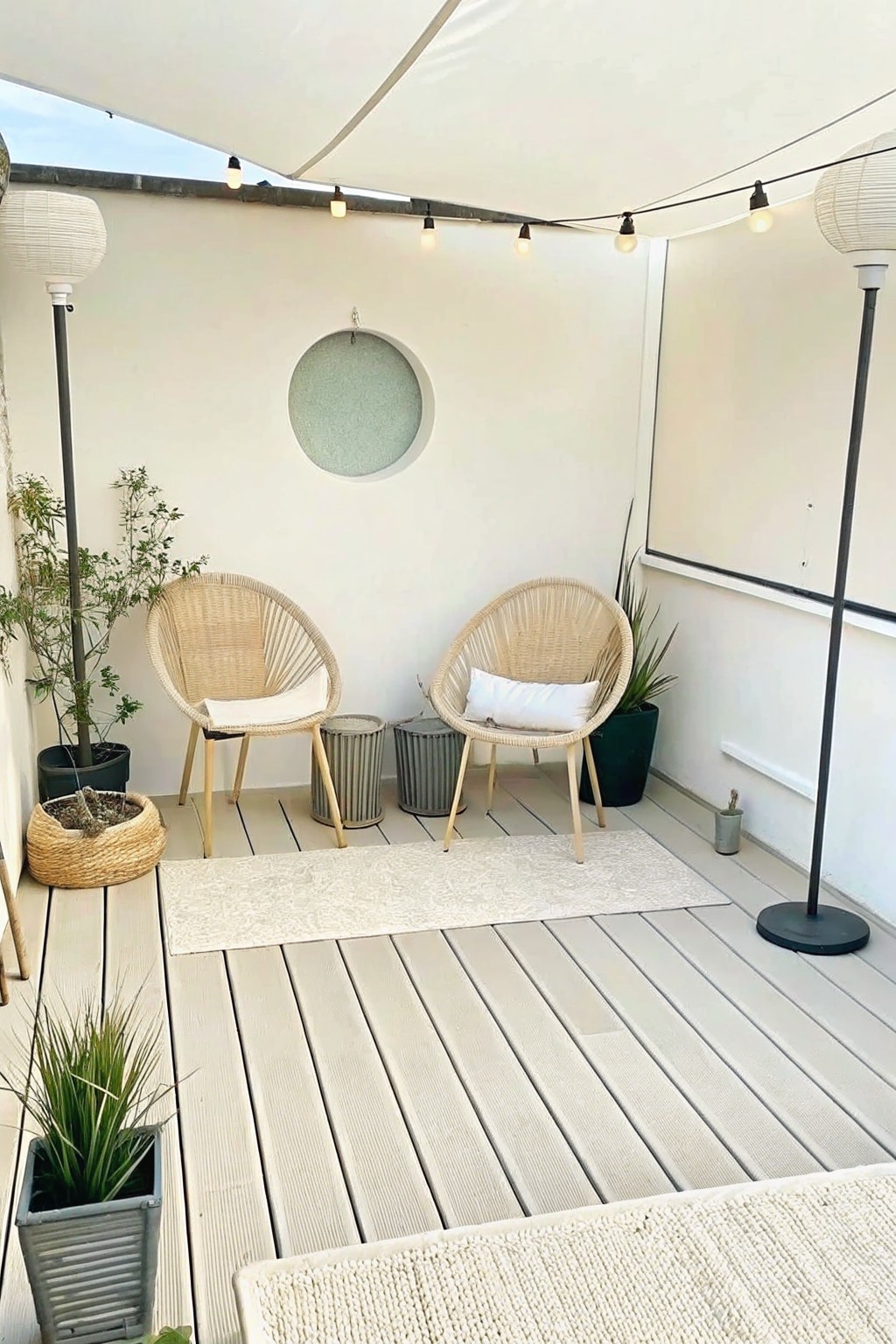 A light-colored deck holds two woven chairs on a patterned rug, with potted plants, string lights, and a white canopy overhead.