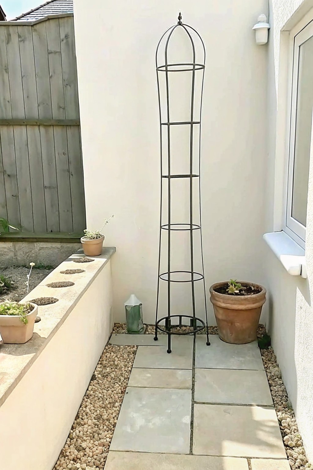 A tall black metal obelisk stands on stone pavers beside a gravel bed with several potted plants along a white wall.