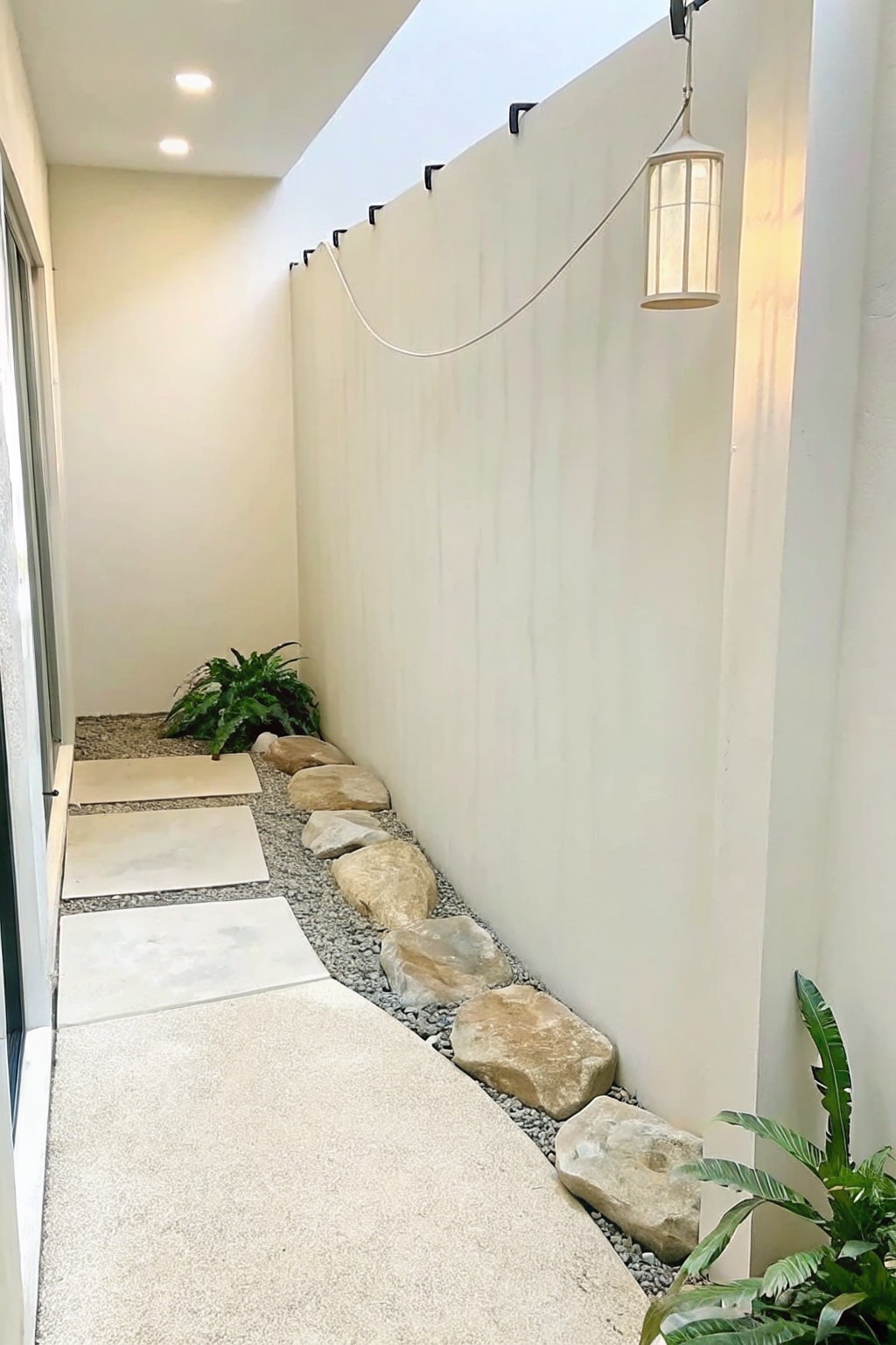 A narrow side yard path with concrete pavers on the left, gravel in the middle, and large boulders lining the right side against a white wall, with a hanging lantern above.