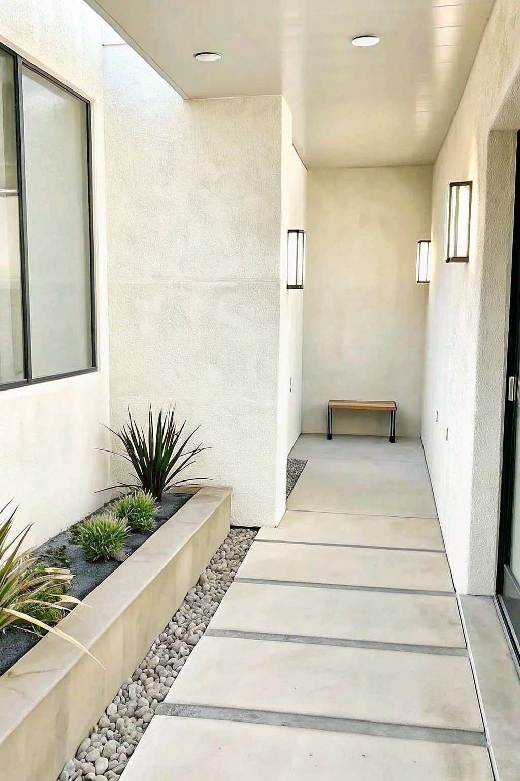 A covered walkway with large concrete pavers runs beside a long planter bed filled with dark gravel, small plants, and a border of light pebbles.