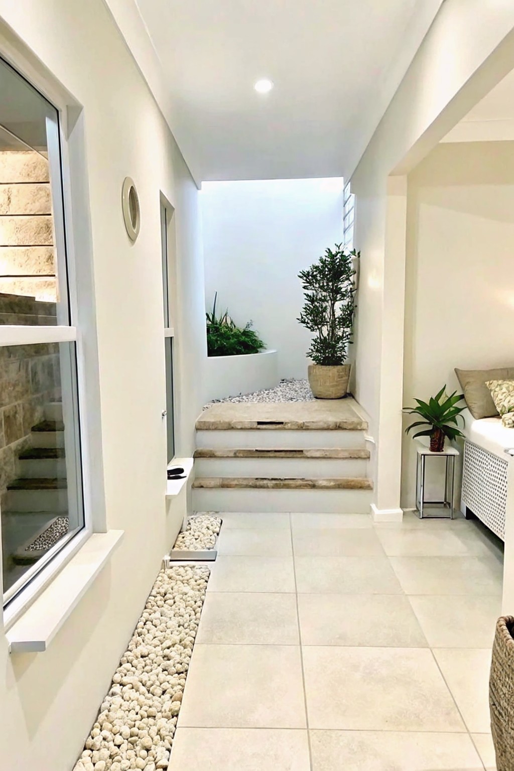 A modern interior hallway with light floor tiles and white gravel beds running along the sides, leading to stone steps and an outdoor courtyard with potted plants.