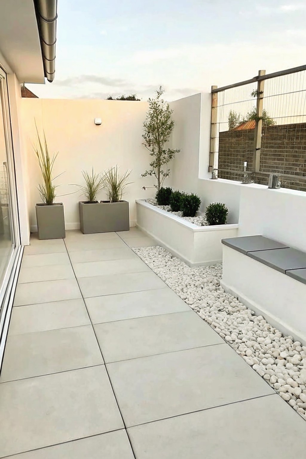 A modern patio with large light tiles, a long white raised planter filled with white gravel and small shrubs, and built-in steps with gray cushions.