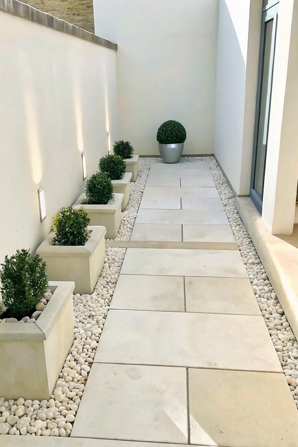 A narrow outdoor walkway lined with large rectangular light stone pavers and bordered on both sides by white gravel, with square planters holding shrubs along the left wall.
