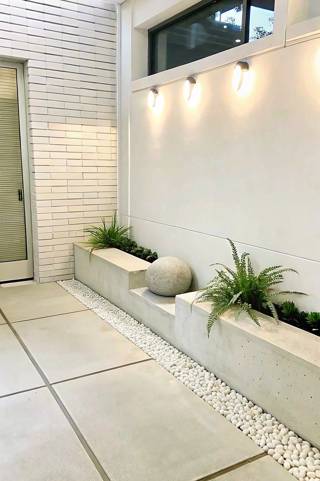 A long concrete planter with ferns and a large gray boulder sits beside white gravel pebbles and light gray tile flooring against a white building wall.