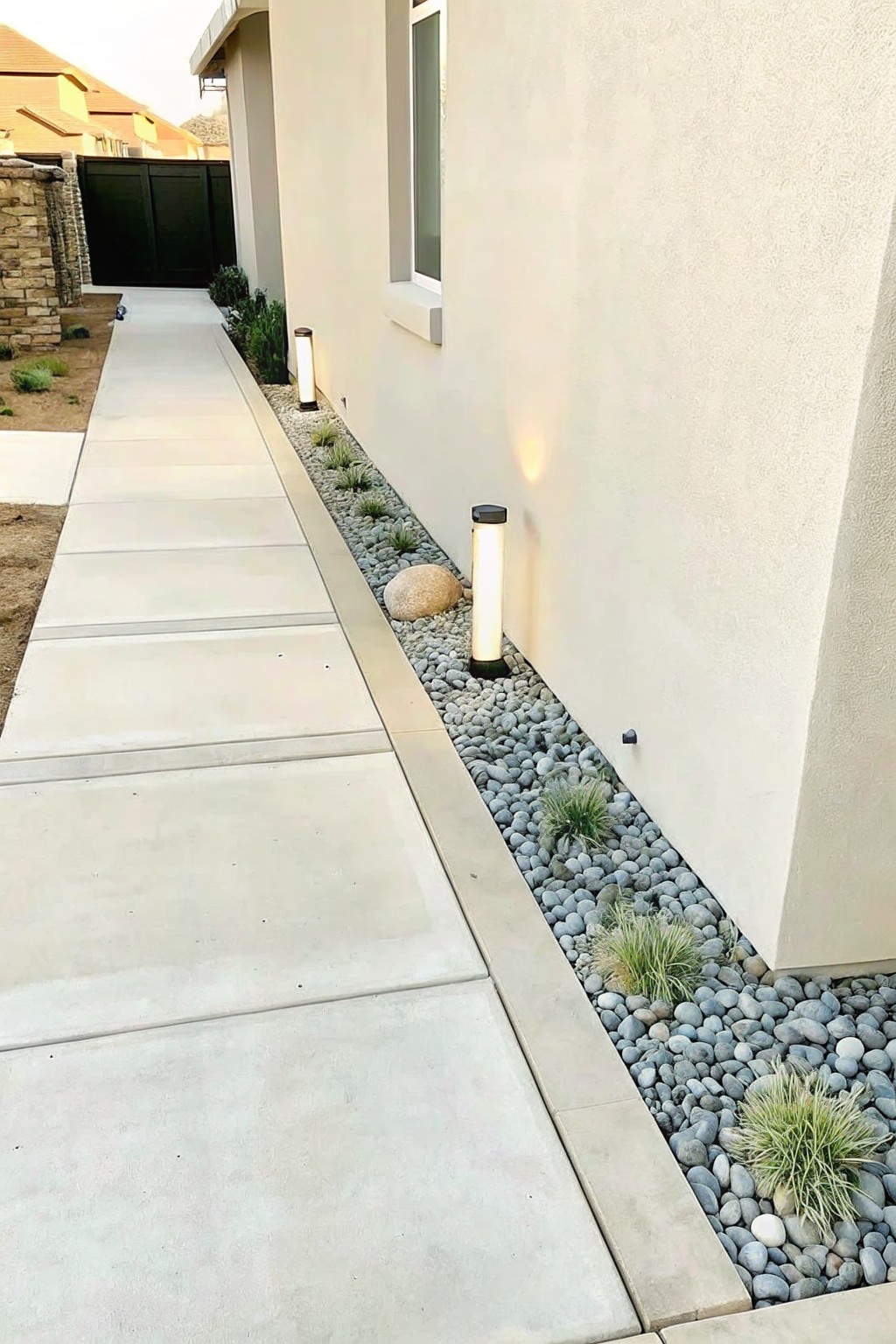 A concrete path runs beside a light-colored house wall with a narrow strip of gray pebbles holding small grasses and two tall cylindrical lights.