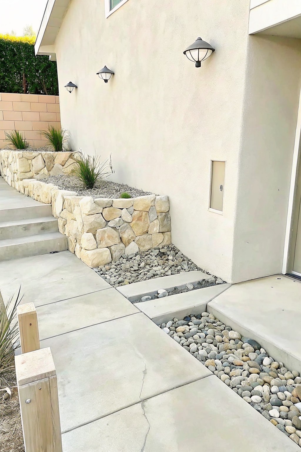 Exterior view of a house showing a stacked stone retaining wall bordering gravel beds beside concrete steps and paths.
