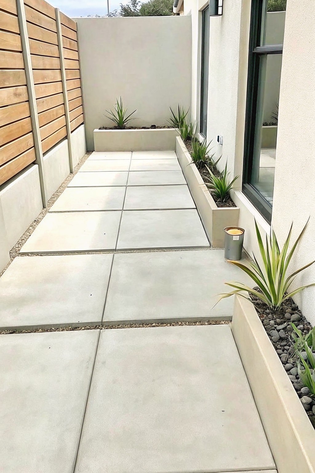 A narrow walkway of large concrete pavers with gravel edges runs beside built-in concrete planters filled with plants, next to a wooden fence and house wall.