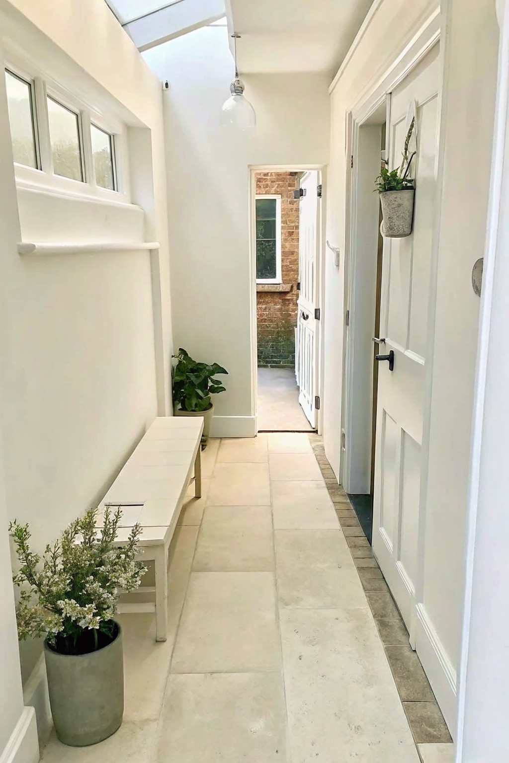 Narrow white hallway with bench, potted plants, hanging greenery, doors, and skylight.