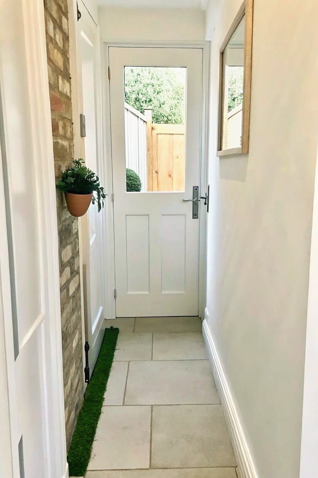Narrow hallway with brick wall, hanging plant, mirror, white door, and tiled floor