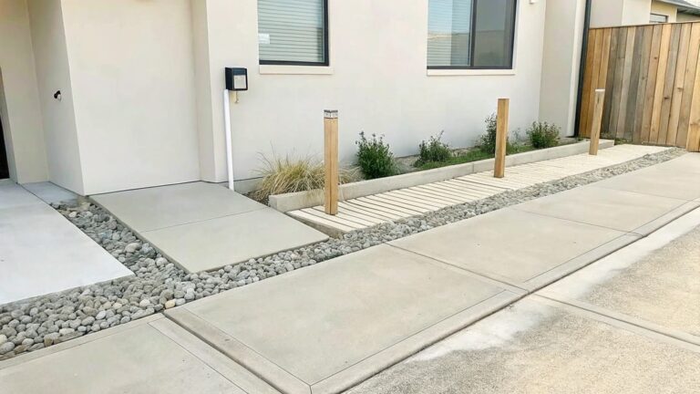 17 Unique Garden Border Ideas Built Around Contrasting Materials A narrow concrete walkway lined with large square pavers set in gray pebbles, running beside a raised garden bed with young plants and leading to a white door.