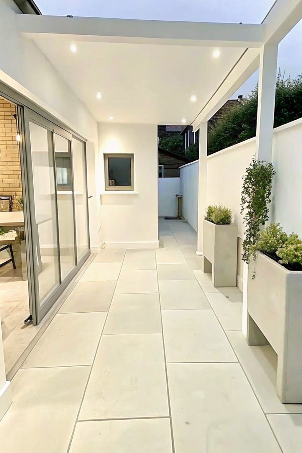 A covered outdoor walkway paved with large light square tiles, white walls on both sides, sliding glass doors on the left, and several rectangular concrete planters holding green plants on the right.